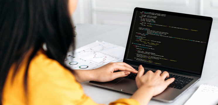 Panoramic over shoulder view female IT-specialist to a laptop with programming code on the screen. Smart professional female programmer working with a software and coding technology, writing codes