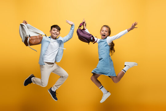 Excited multiethnic kids with backpacks jumping on yellow background.