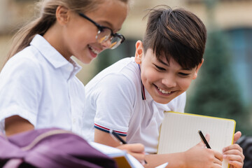 Positive asian schoolboy holding pen and notebook near friend outdoors.