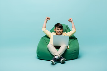 Asian schoolkid showing yes gesture while looking at laptop on beanbag chair on blue background.