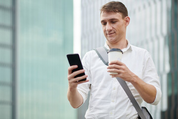 Young entrepreneur drinking take-out coffeee and ordering car via taxi application on smartphone