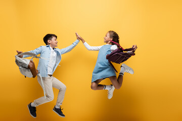 Fototapeta na wymiar Side view of multiethnic schoolkids with backpacks giving high five while jumping on yellow background.