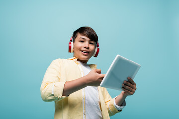 Low angle view of smiling asian schoolboy in headphones holding digital tablet isolated on blue.