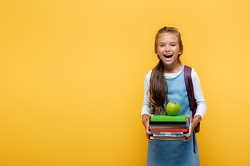 Cheerful pupil holding books and apple isolated on yellow.