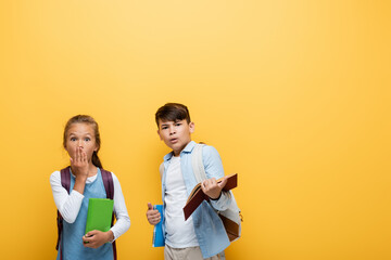 Shocked interracial pupils with books looking at camera isolated on yellow.