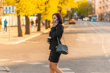 Young business woman talking on the phone holding coffee paper cup,crossing street blurred background. Fashion business photo of beautiful girl in black suite with phone and cup of coffee.Side view.