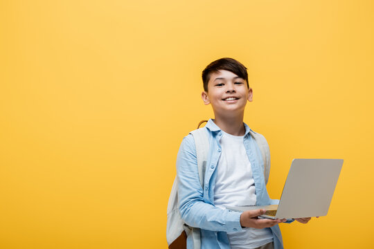 Smiling asian schoolboy holding laptop isolated on yellow.