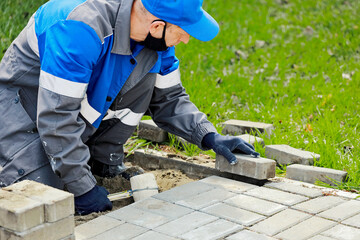 Bricklayer in work clothes sits on sidewalk and lays out paving slabs. Sight of working man in open air. Professional builder makes arrangement of territory summer day. Real scene.