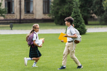 Obraz premium Smiling multiethnic classmates with notebooks running on lawn in park.