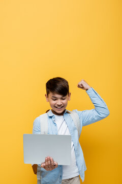 Excited asian schoolkid showing yes gesture and holding laptop isolated on yellow.