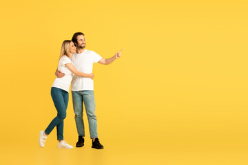 Fototapeta na wymiar Glad happy millennial caucasian man and lady in white t-shirts hugging and pointing finger at empty space