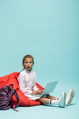 Shocked schoolgirl using laptop on beanbag chair on blue background.