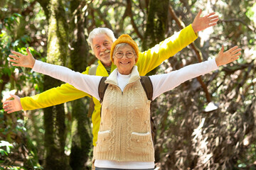 Cheerful caucasian senior couple with backpack standing in the forest with outstretched arms...