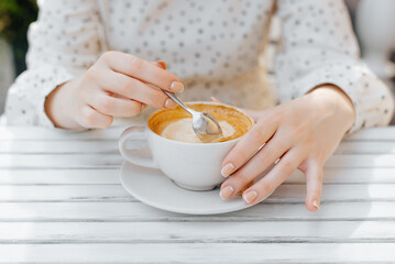 Close-up of female hands stirring cappuccino with spoon in white cup while sitting at table in street cafe, outdoors on summer day. Coffee time, morning breakfast concept