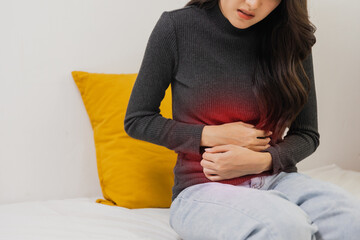 Young woman suffering from strong abdominal pain while sitting on bed at home