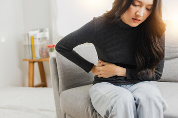 Naklejka premium Young woman suffering from strong abdominal pain while sitting on sofa at home