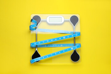 Electronic scales and measuring tape on yellow background, top view