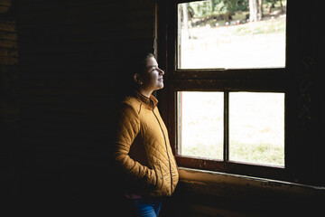 Vertical image of a smiling woman looking out the window with a mustard-colored jacket and her hands inside the jacket pockets. 