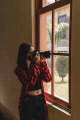 Vertical portrait of a teenage girl inside a building, taking a picture with her camera through a window. 
