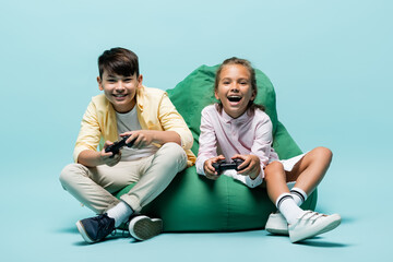 KYIV, UKRAINE - JULY 2, 2021: Happy multiethnic preteen kids playing video game on beanbag chair on blue background.