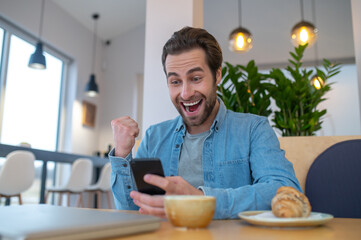 Fototapeta na wymiar Enthusiastic man looking at smartphone sitting at table