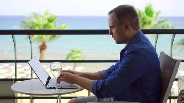 Businessman working on laptop at tropical beach resort. Male freelancer drinking wine from wineglass