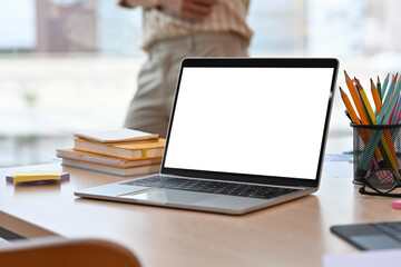 Workplace desk with a laptop with a white blank screen, note books, pencils and a person in a background, for business and technology concept.