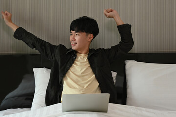 A portrait of a handsome Asian man lying on bed, stretching  while working on a laptop. For...