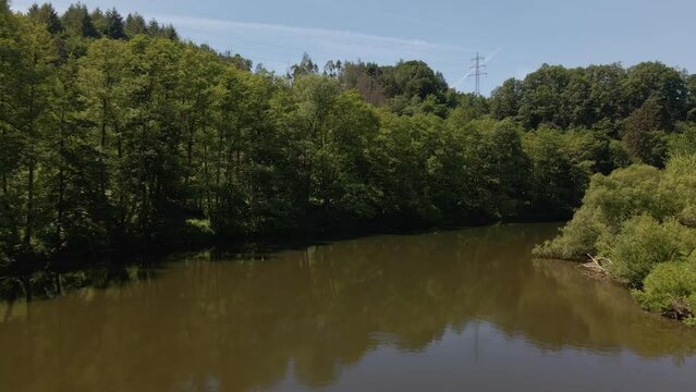 Wide angle drone footage following a meandering brown river in between lush green banks covered by thick deciduous forests and bushland. Aerial flyover revealing a small bridge behind a curve
