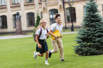Obraz premium Smiling multiethnic schoolkids with notebooks running on lawn in park.