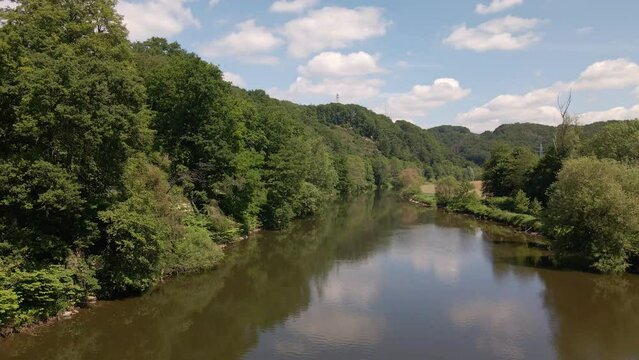 Calm, brown river surrounded with lush and leafy riverbanks underneath a blue sky with cumulus clouds. Aerial pull back shot