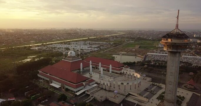 Circle drone shot of the great Mosque of Central Java (MAJT) in suset time. Th sun is orange and the weather is cloudy. It have some pillars and some umbrela that is closed and A big tower
