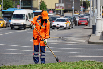 Woman worker in orange uniform with a broom sweeps the road on cars background. Street cleaning in summer city