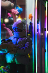 Girl with a dog toy against the background of a showcase with toys with neon illumination. Smiling girl in warm clothes: a jacket, a hat, a scarf at night. Holidays theme