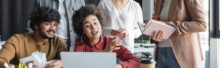 african american woman talking near laptop and indian colleague in advertising agency, banner.