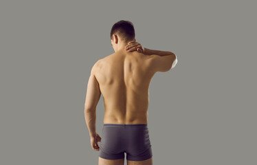 Fototapeta na wymiar Rear view of naked sporty man in underwear isolated on grey studio background suffer from neck ache. Pain in upper back. Sexy toned guy in briefs struggle with backache or muscular spasm.