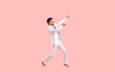Fototapeta na wymiar Cheerful funny and active young man in suit having fun isolated on pastel pink background. Stylish man in white formal suit, sneakers and glasses cheerfully waving his arms. Full length. Copy space.