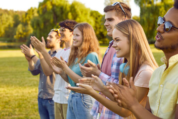 Group of joyful contented multiracial young people applaud and cheer together in park. Side view of...