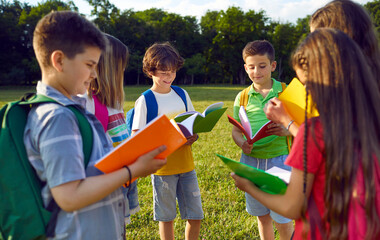 Group of elementary school students read books together standing in circle in park near school. Smiling preteen boys and girls with colored books and with backpacks study together in park on sunny day