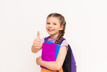 A schoolgirl with a satchel and books gives a thumbs up on a white isolated background and smiles. Preparatory courses for schoolchildren.