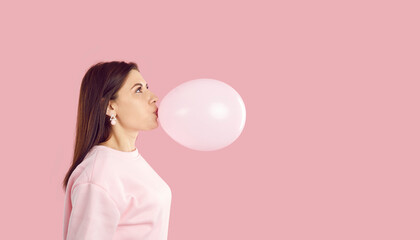 Side view of young woman blowing up pink balloon. Profile view of happy brunette girl inflating balloon on pink copy space studio background. Birthday party and fun concept