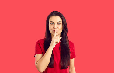 Portrait of young woman on red studio background hold finger at lips make shush sign. Millennial...