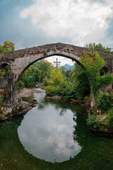 Fototapeta na wymiar Famous Roman bridge over a lake, in Cangas de Onís, Asturias, with a lot of vegetation and a cross hanging over it. 