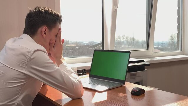 A frustrated Caucasian man sits at a desk in front of a computer with a green screen in the office and rubs his face with his hands. Workaholic, hard work, career ladder, earning money. High quality
