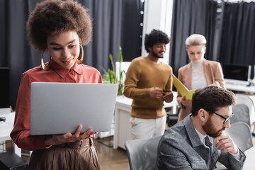 positive african american woman looking at laptop near multiethnic colleagues working in ad agency.