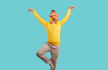 Cheerful young man in rubber rooster mask shows funny dance moves on turquoise background. Man with a chicken mask on his head, wearing yellow shirt and pants having fun on colored background. © Studio Romantic