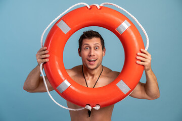Male beach rescuer. Surprised bearded man holding inflated lifebuoy with wide opened mouth and being ready to rescue you isolated over blue background