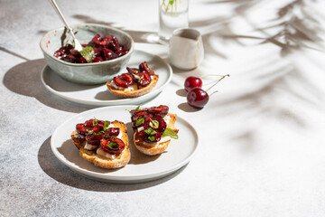 Toasts with soft cheese and baked with herbs sweet cherry berries on white concrete background. Roasted cherries. Delicious breakfast idea.