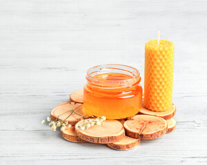 Glass jar with honey, beeswax candle, white wildflower on a wooden stand on a white wooden background, front view, close-up, copy space. Alternative medicine, healthy eating, vegetarianism. - obrazy, fototapety, plakaty