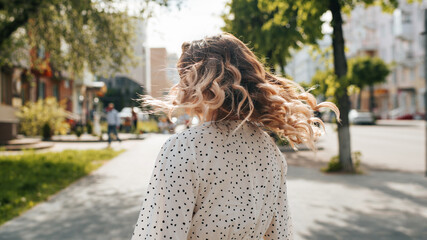 Fototapeta na wymiar Stylish woman with curly hairstyle is spinning on city street, back view. Unrecognizable free carefree lady, summer lifestyle mood. Selective focus on dress with polka dots, hair blur in motion